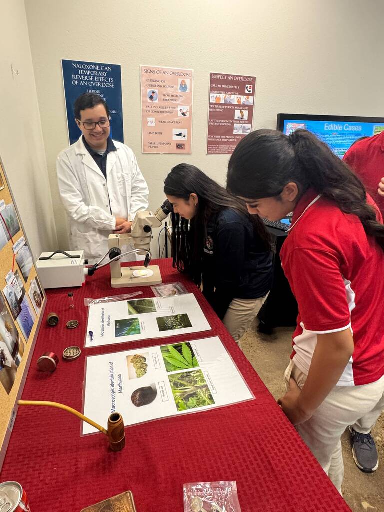 A man in a white lab coat stands near a microscope while two students in red shirts observe.