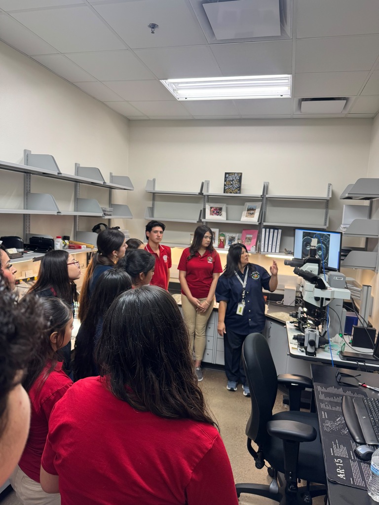 A group of people wearing red shirts stand in a lab, looking at a microscope on a desk.