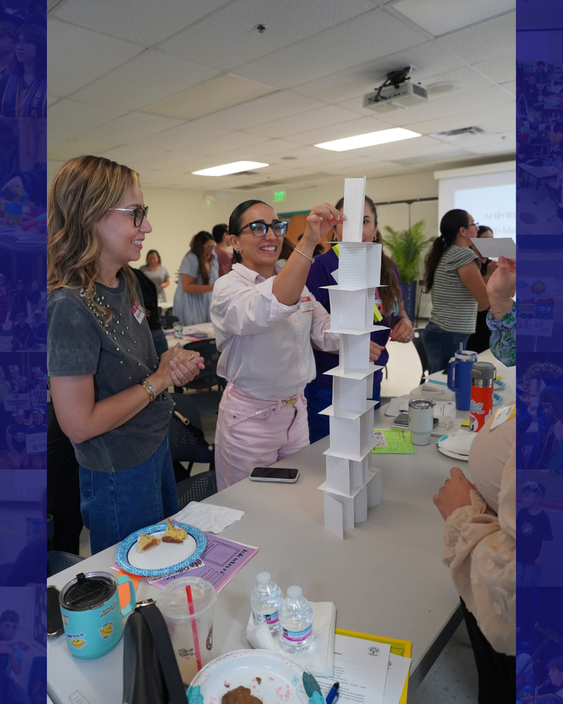 Two women attempt to build a tower of cards in a room with other people and objects.