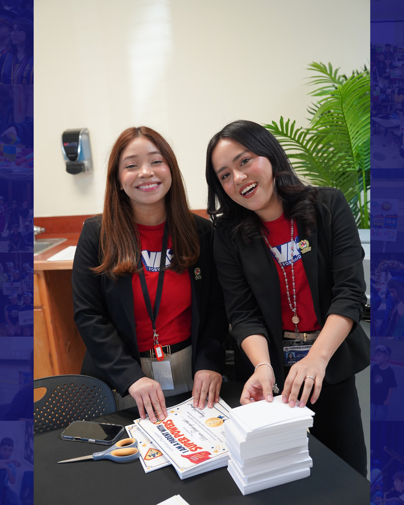 Two women wearing red shirts and black blazers stand behind a table with a stack of white cards.