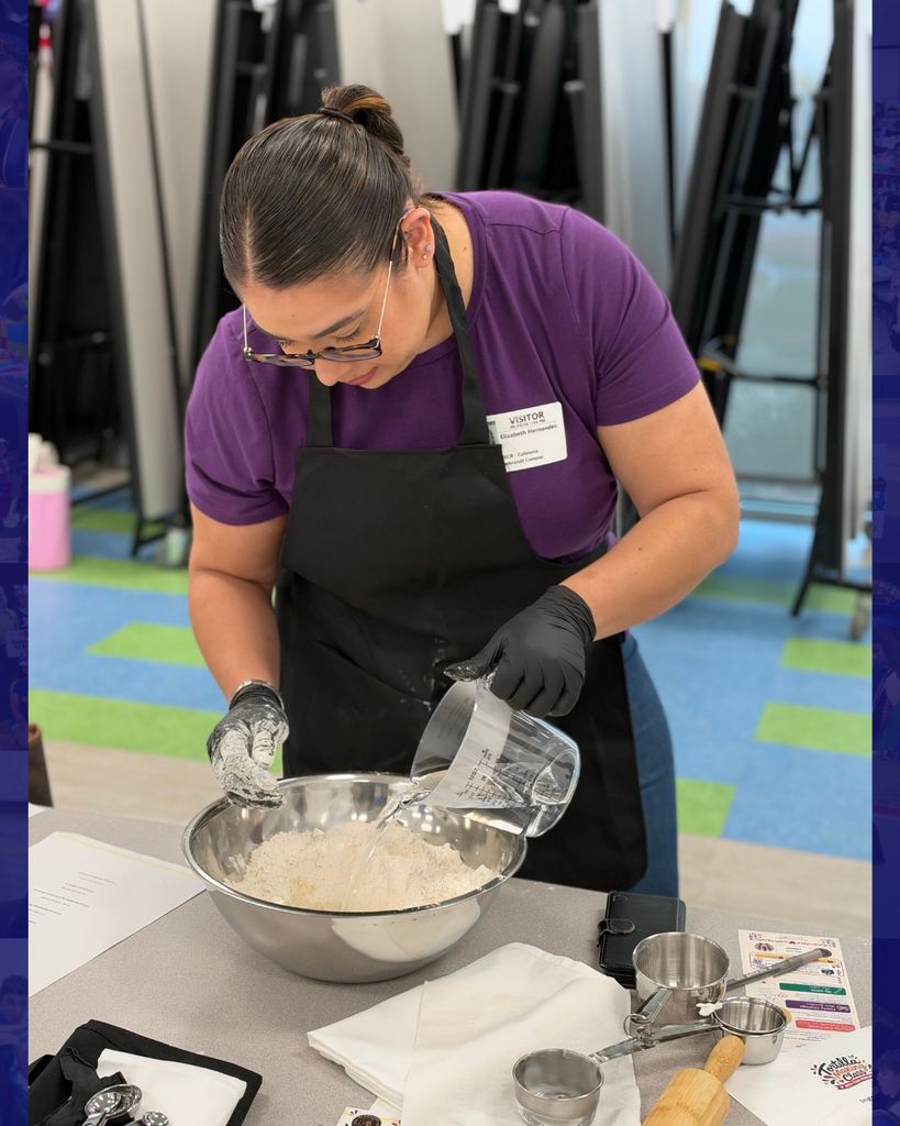Woman in purple shirt and glasses wearing black apron, pouring ingredients into a bowl for baking.