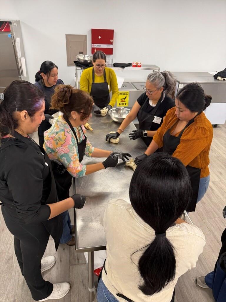 Women in aprons prepare food in a kitchen. A woman leads the group around a large table.