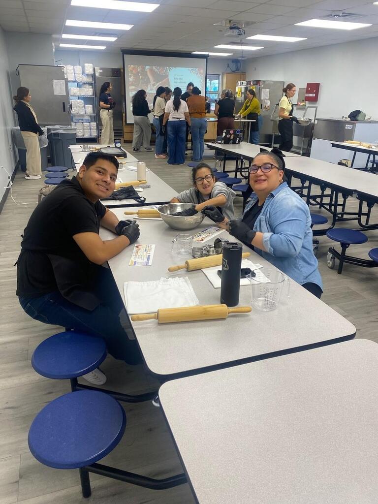 Three people at a table with a bowl, rolling pins, and cups. Another group is standing in the background.