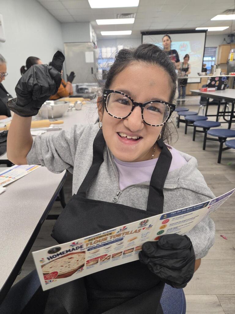 Girl wearing black gloves and glasses, holding a book, smiling, and sitting at a table.