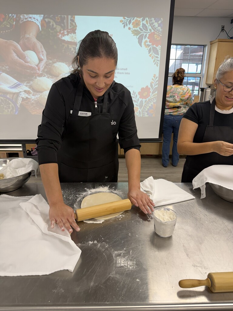 A woman in a black apron rolls dough on a counter. Another woman stands behind her. A projector screen shows images of hands rolling dough.