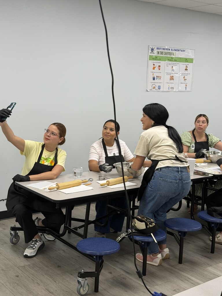 Five people in aprons around a table. One woman holds a phone and others are seated. A white wall with a green sign is behind them.