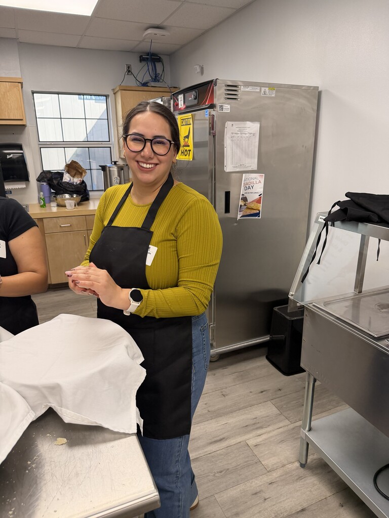 A smiling woman in a yellow sweater and glasses stands in a kitchen with a stainless-steel fridge.