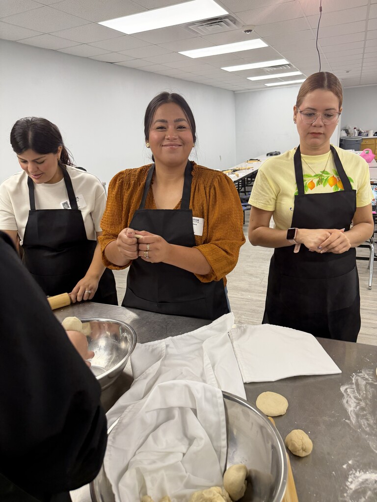 Three women in aprons roll dough in a kitchen. Bowls and dough balls on a table.