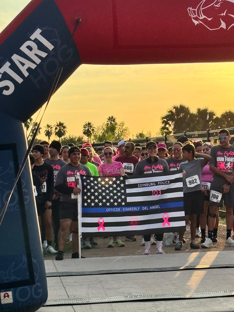 A group of adults in T-shirts, hats, and sunglasses stand behind a blue arch. They hold a flag with the American flag and a ribbon.