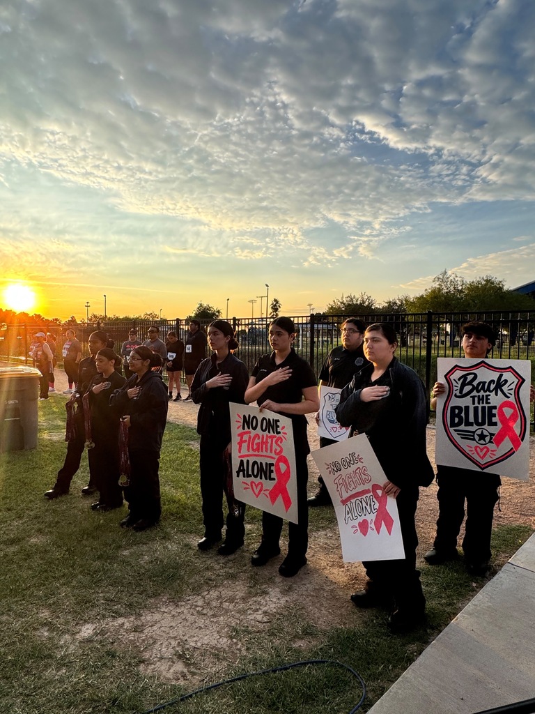 People hold signs outdoors, standing on grass. One sign reads "Back the Blue." The sun sets behind them.