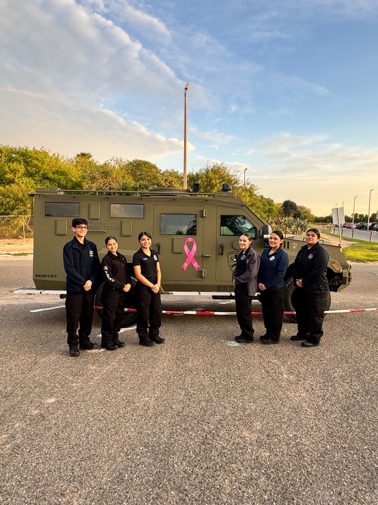 Six people stand in front of a green vehicle with a pink ribbon. Behind them are trees and a clear sky.
