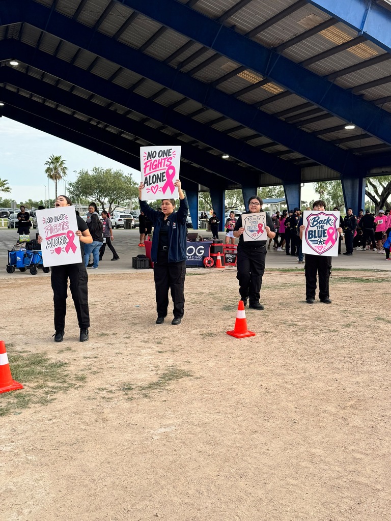 Four adults hold signs in a dirt field with orange cones and blue roof overhead.