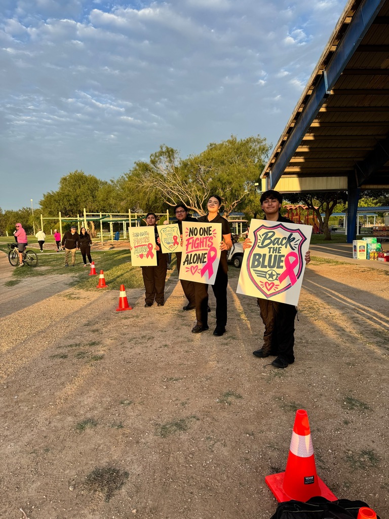 People holding signs with messages about breast cancer awareness standing in a dirt area. Traffic cones and a bridge in the background.