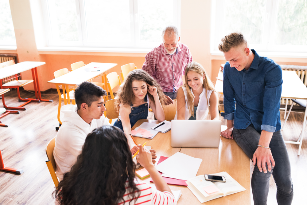 students and teacher around a desk looking at school work and laptop