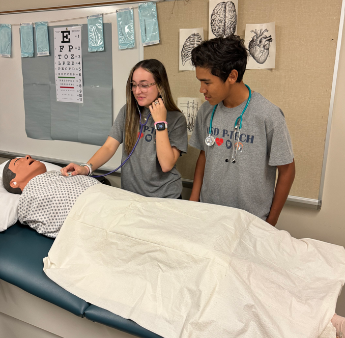 Two Medical PTECH students listening to heart with a stethoscope  on a  mannequin laying on a  medical table