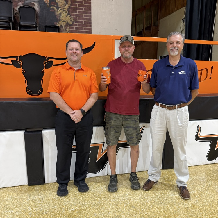 Three men standing indoors in a gymnasium with an orange Longhorn mascot mural. From left: a man in an orange polo and black pants, a man in a maroon t-shirt and camo shorts holding two orange mugs, and a man in a navy polo and white pants.