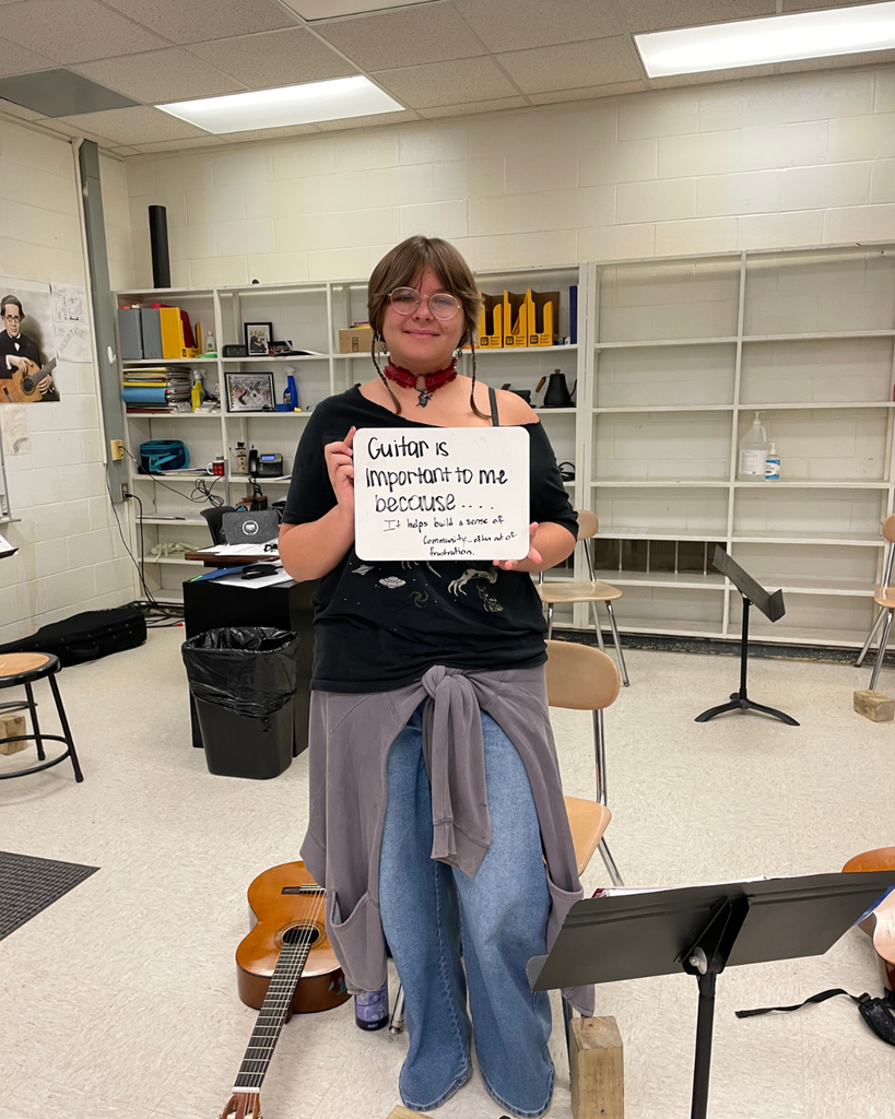 The image is a high school student holding a sign that says "guitar is important to me because...it helps build a sense of community...often out of frustration."