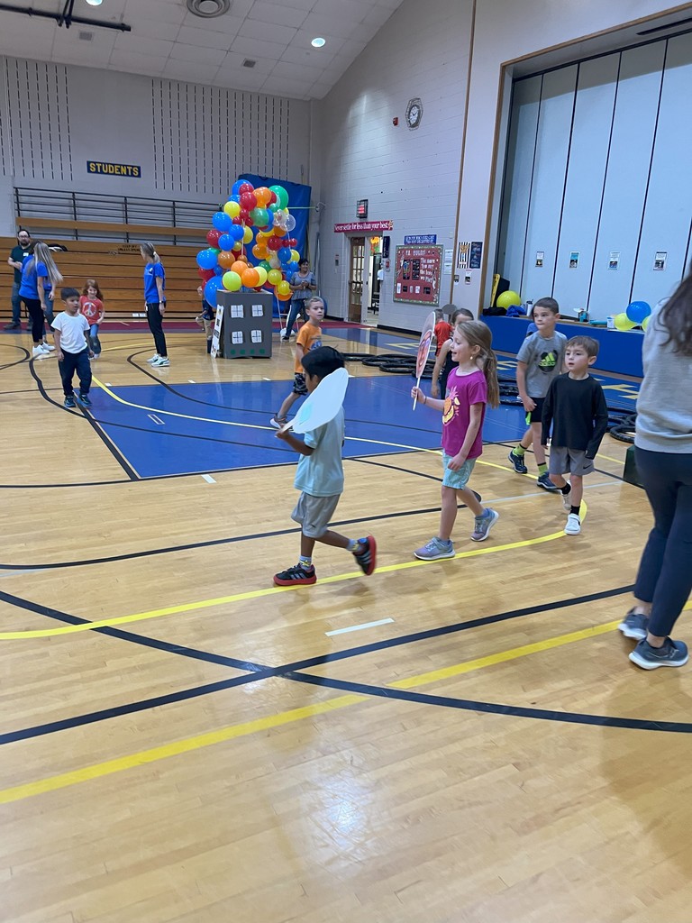 Children playing in a colorful, gymnasium-like indoor space with balloons, basketball hoops, and activity stations.