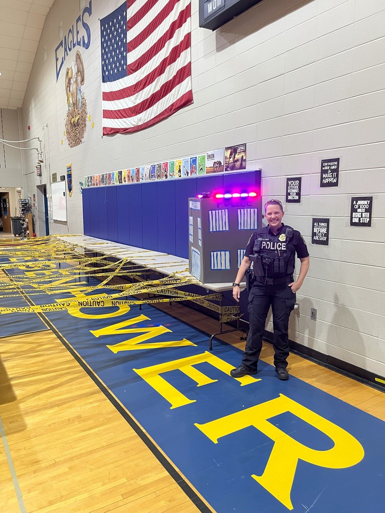 A police officer stands in a school gymnasium, surrounded by a large American flag, lockers, and yellow caution tape on the floor.