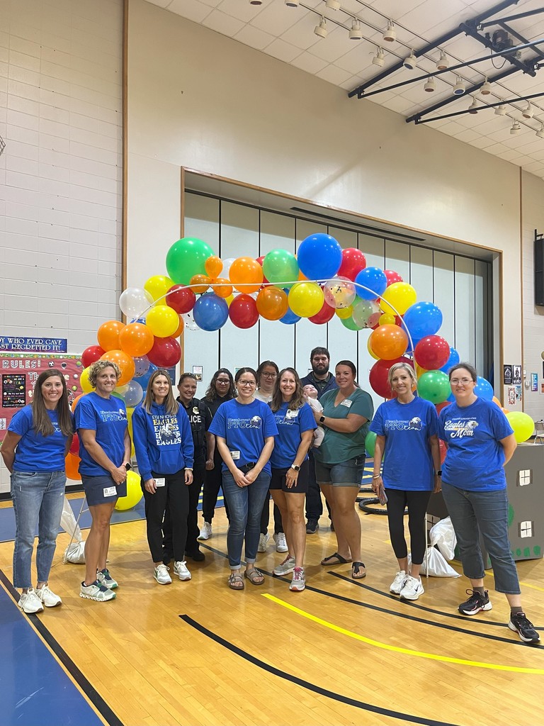A group of people wearing matching blue shirts standing in front of a colorful balloon arch in a gymnasium.
