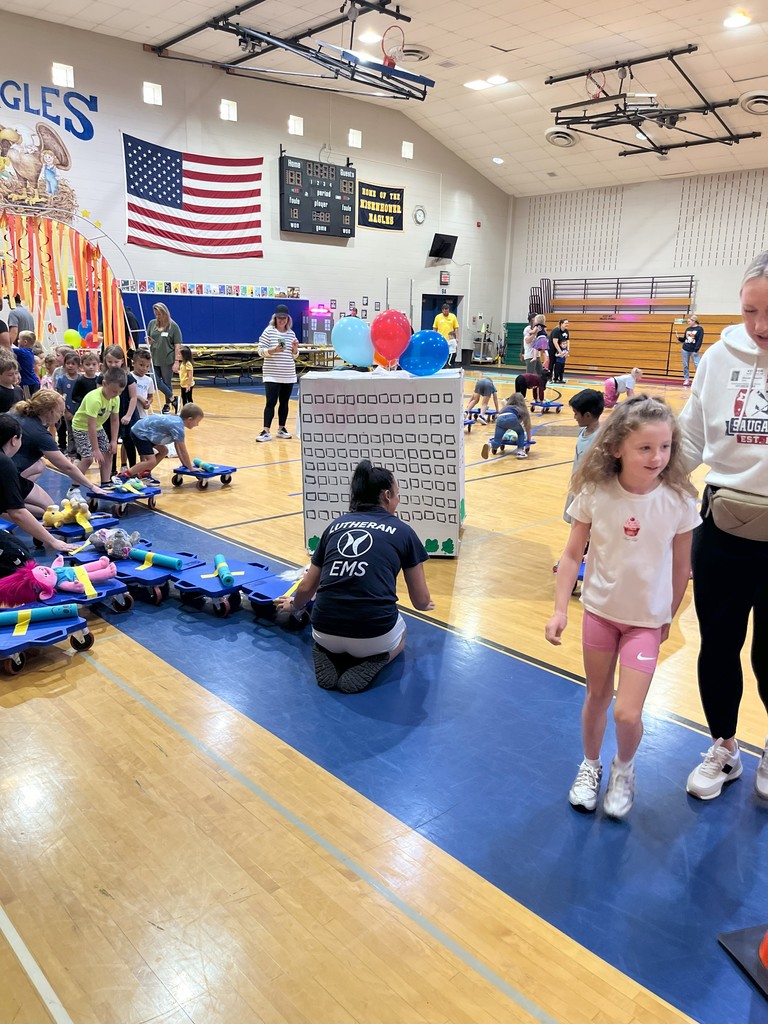 A crowded school gymnasium with students playing on scooters and floor mats, an American flag on the wall, and a scoreboard visible.