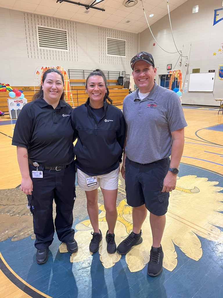 Three smiling people in uniform standing in a school gymnasium with sports equipment visible in the background.
