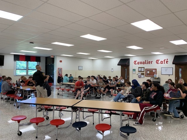 Students listen to a police officer speak in a cafeteria.