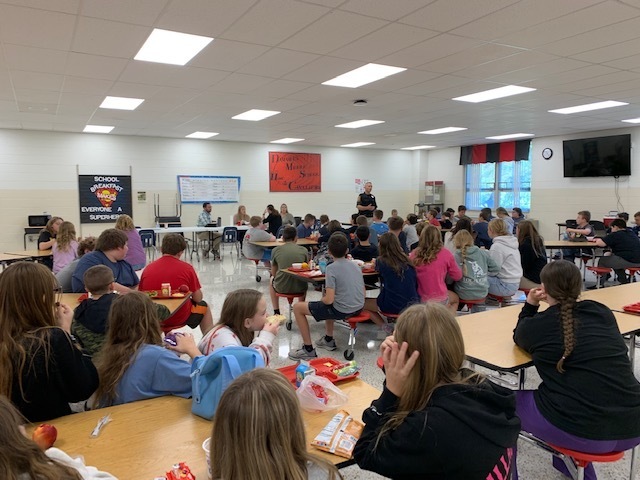 Students listen to a police officer speak in a cafeteria.