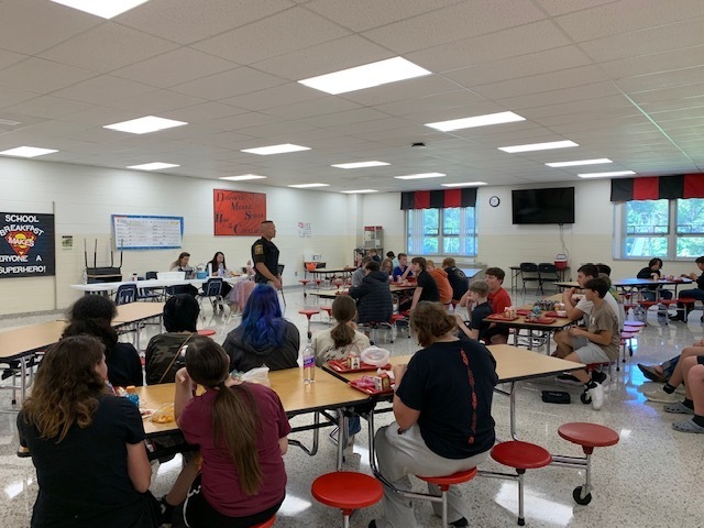 Students listen to a police officer speak in a cafeteria.