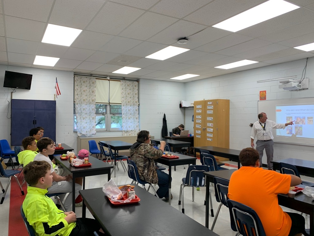Students listen to a speaker while eating lunch in a classroom.