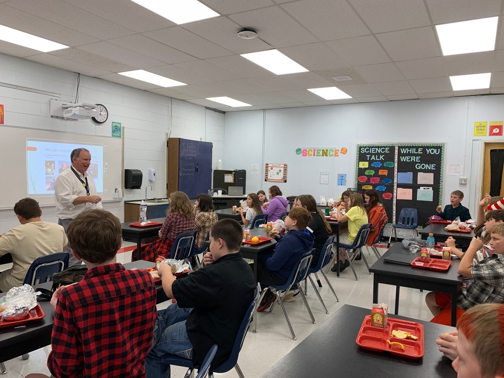 Students listen to a speaker while eating lunch in a classroom.
