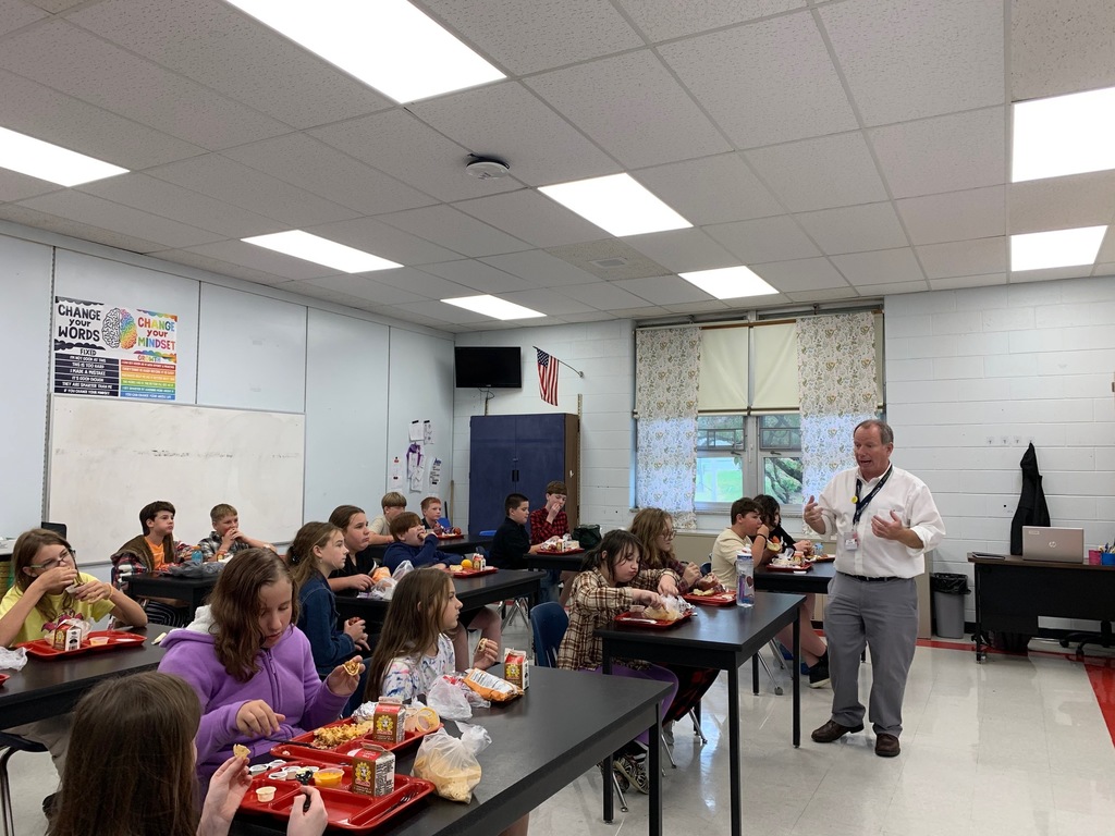 Students listen to a speaker while eating lunch in a classroom.
