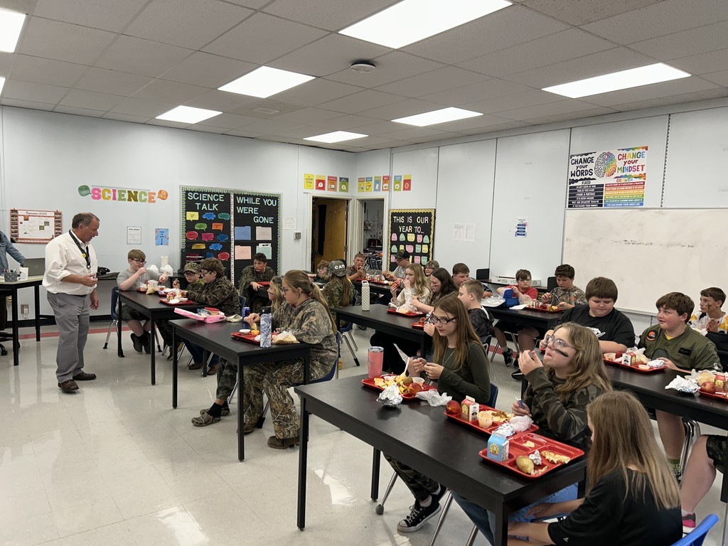 Students listen to a speaker while eating lunch in a classroom.