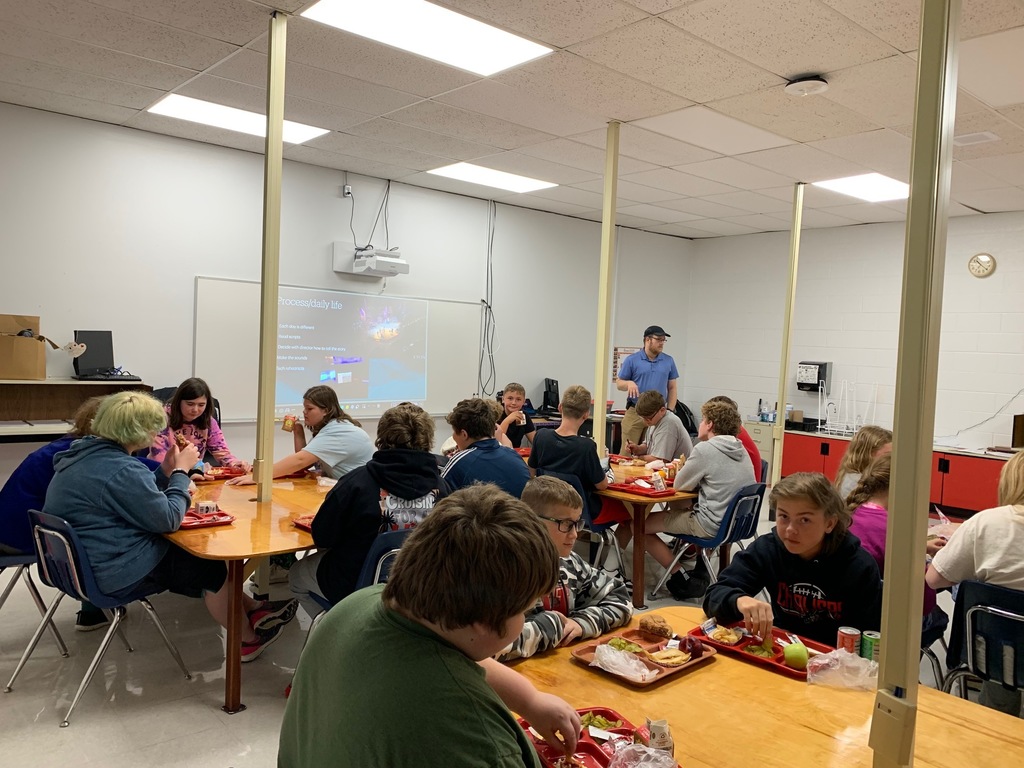 Students eat lunch while a man talks with them.