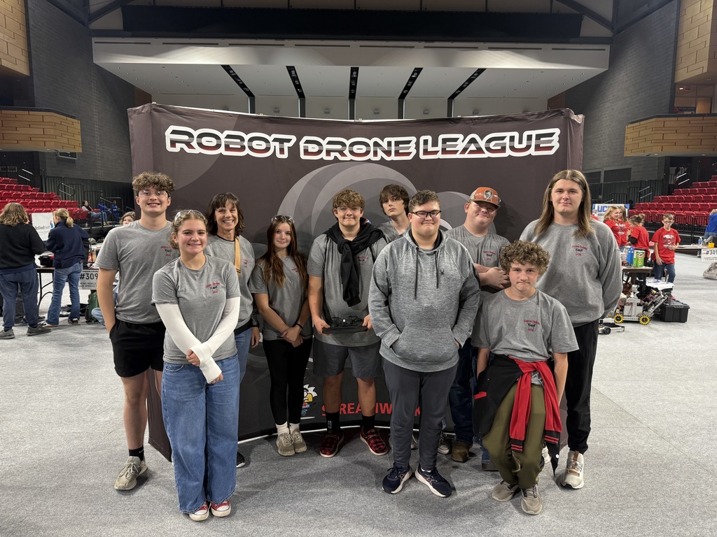 A robotics team and the coaches pose in front of a Robot Drone League poster.