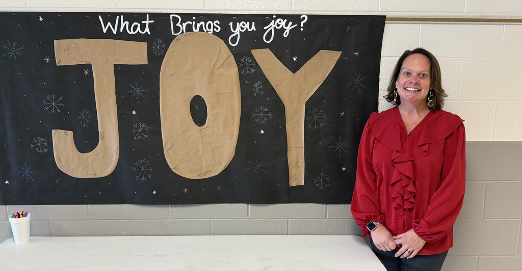 A teacher stands next to a bulletin board that says, "What Brings You Joy?"