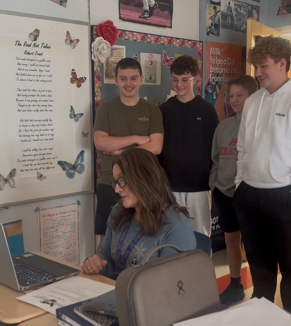 A teacher looks at her computer as four students look over her shoulder.