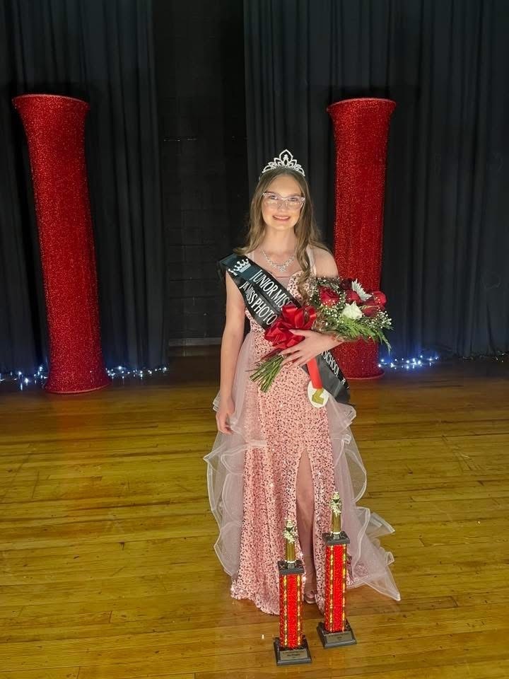 A middle school girl wearing a pageant dress poses on a stage with flowers and trophies.