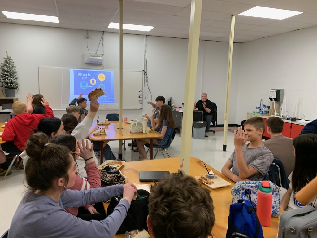 Students listen to a presenter in a classroom while they eat lunch.