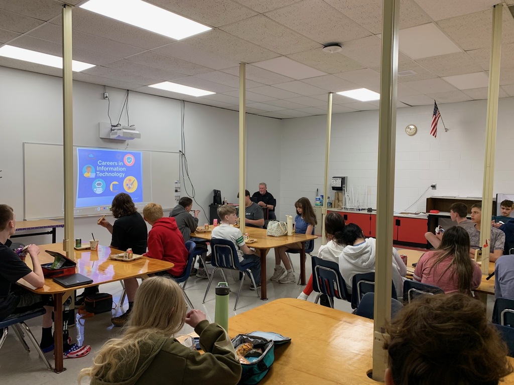Students listen to a presenter in a classroom while they eat lunch.