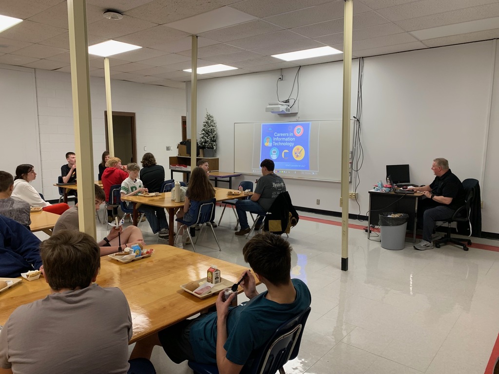 Students listen to a presenter in a classroom while they eat lunch.