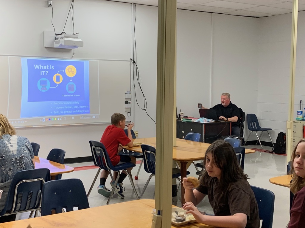 Students listen to a presenter in a classroom while they eat lunch.