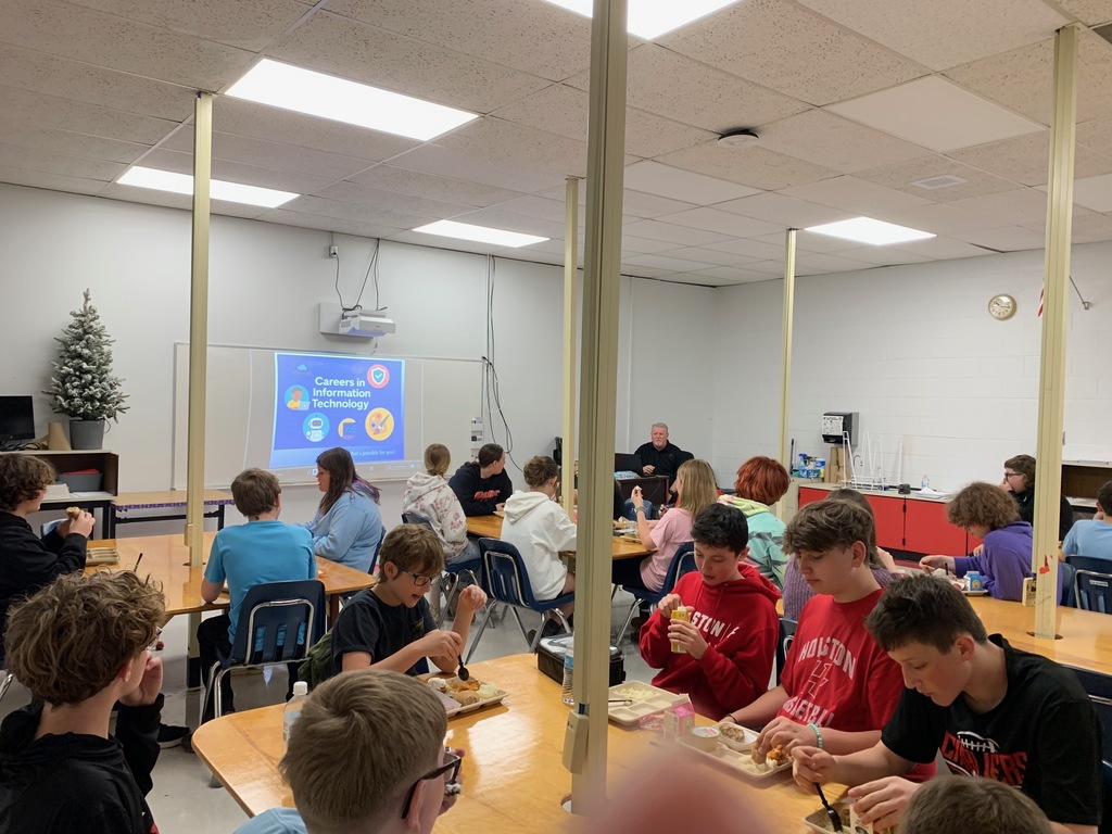 Students listen to a presenter in a classroom while they eat lunch.