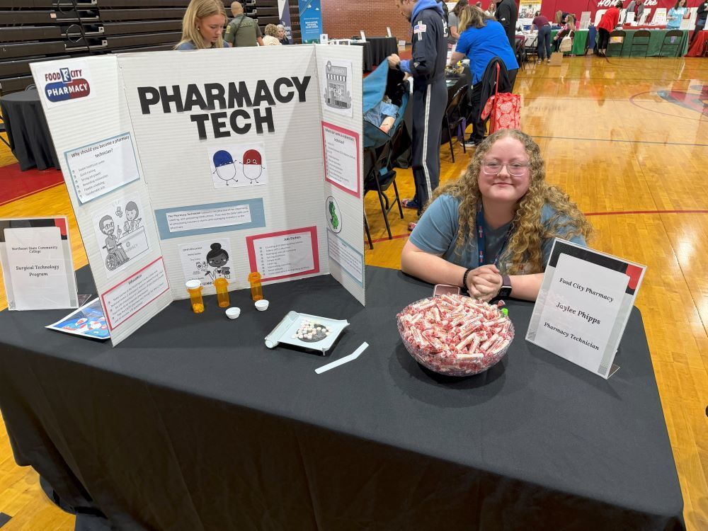 A woman sits at a table with a Pharmacy Tech board and candy.