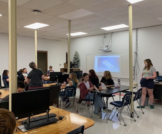 Three students and a teacher talk with a group of younger students in a classroom.
