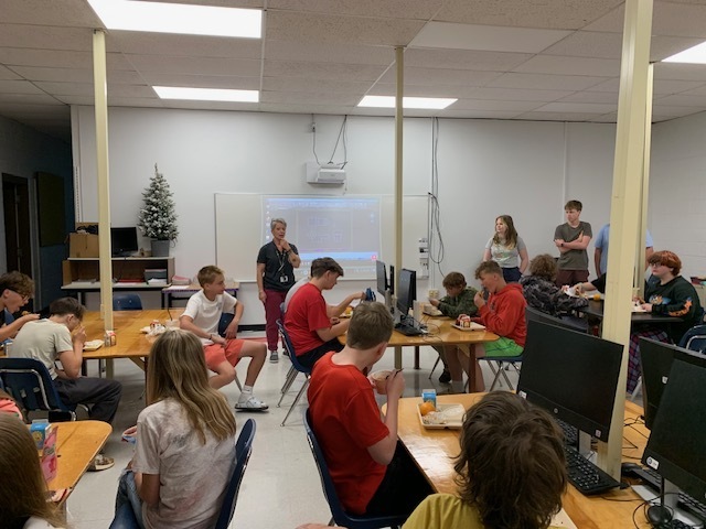 Three students and a teacher talk with a group of younger students in a classroom.