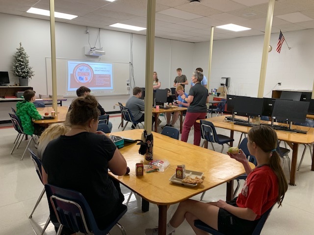 Three students and a teacher talk with a group of younger students in a classroom.