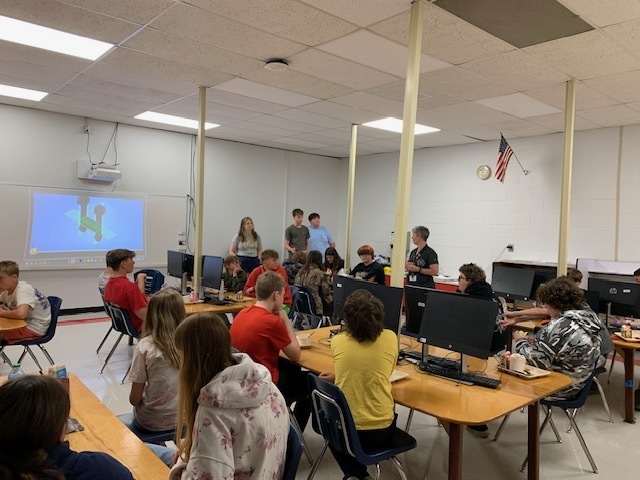 Three students and a teacher talk with a group of younger students in a classroom.
