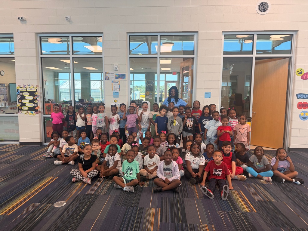 A large group of kindergarten students pose together in a bright school hallway with a staff member standing behind them. The children are smiling and gathered in several rows, with some standing and others sitting on the floor. The group is celebrating Custodian Appreciation Day and recognizing the important work custodians do to care for their school.
