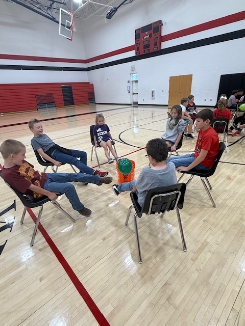 Students playing a teamwork game sitting in chairs and trying to kick a balloon into a bucket.